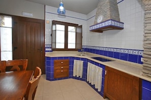 Una cocina con chimenea de piedra, azulejos blancos y azules, armarios de madera, un lavamanos y una mesa de comedor con sillas.