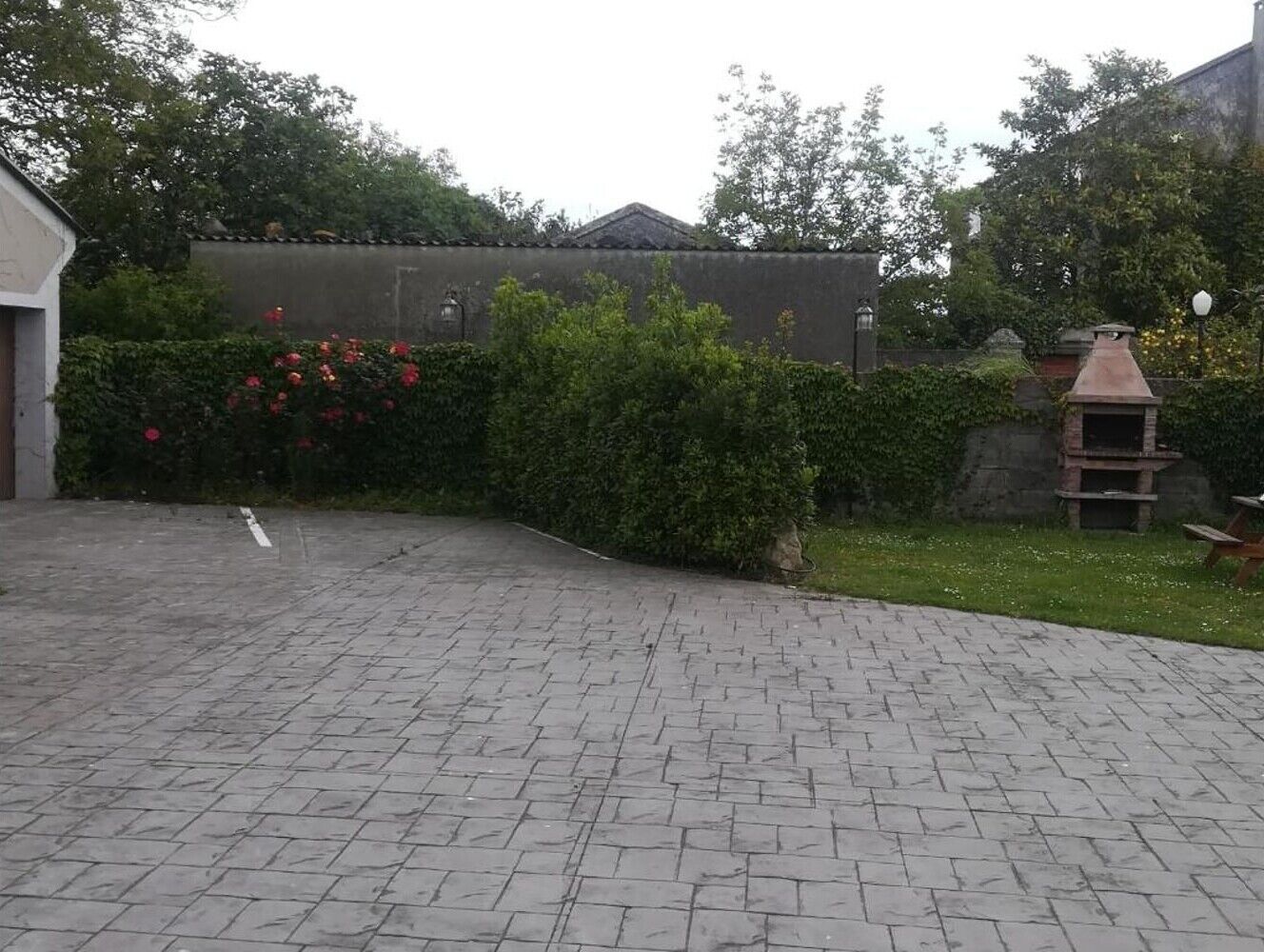 A paved courtyard with a brick floor, a small barbecue grill, and a hedge with red flowers.