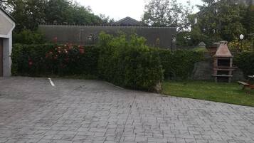 A paved courtyard with a brick floor, a small barbecue grill, and a hedge with red flowers.