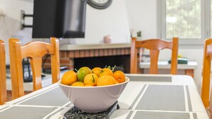 Een eettafel met een fruitschaal, houten stoelen, een televisie en een klok aan de muur.