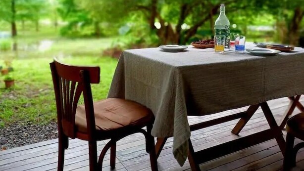 Una terraza de madera al aire libre con una mesa preparada para dos, rodeada de exuberante follaje y árboles.
