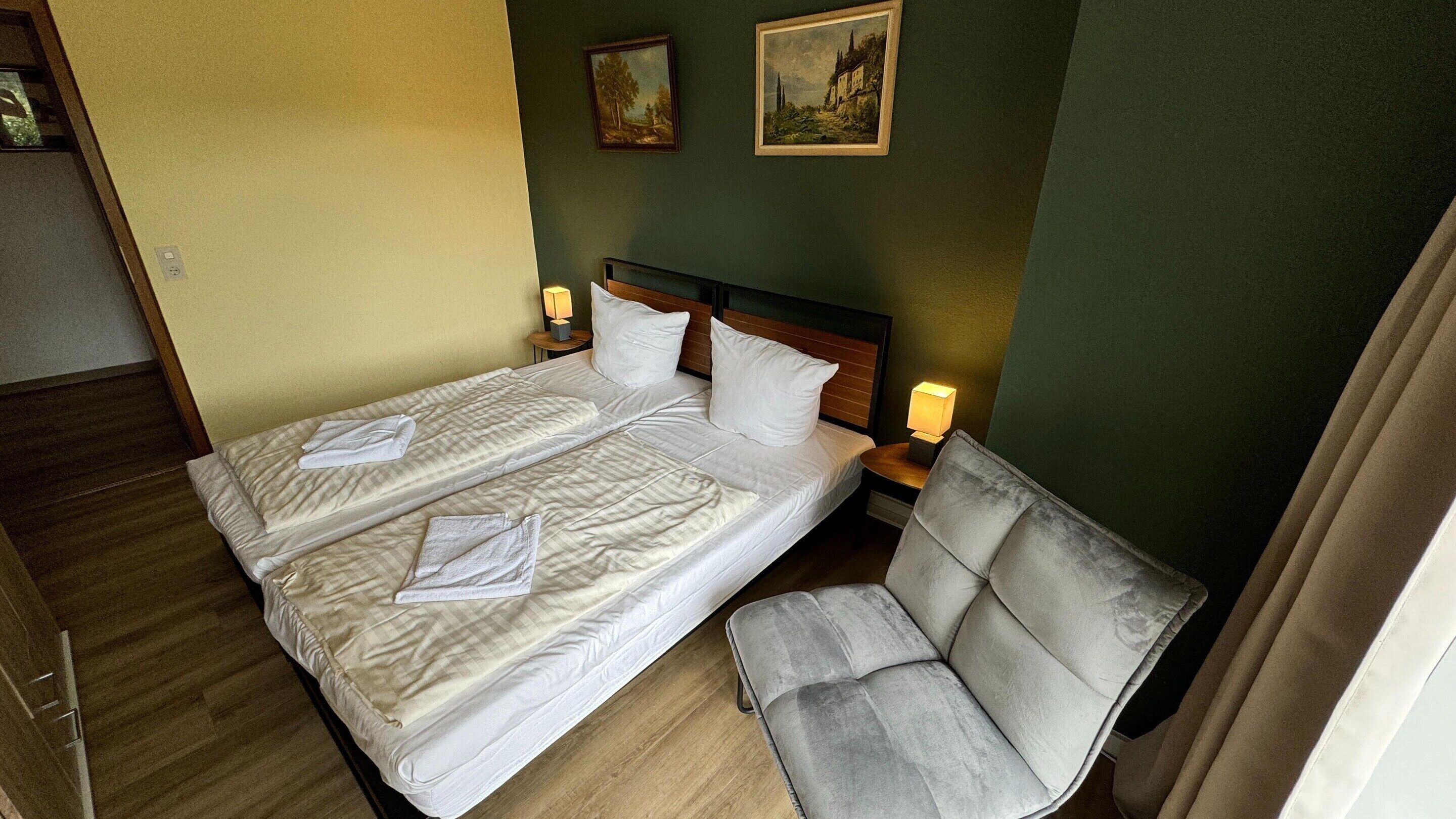 A hotel room with a double bed, two pillows, a bedside table with a lamp, a chair, and two paintings on the wall.