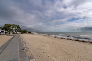 En strand med en asfalterad strandpromenad, träd och havet.