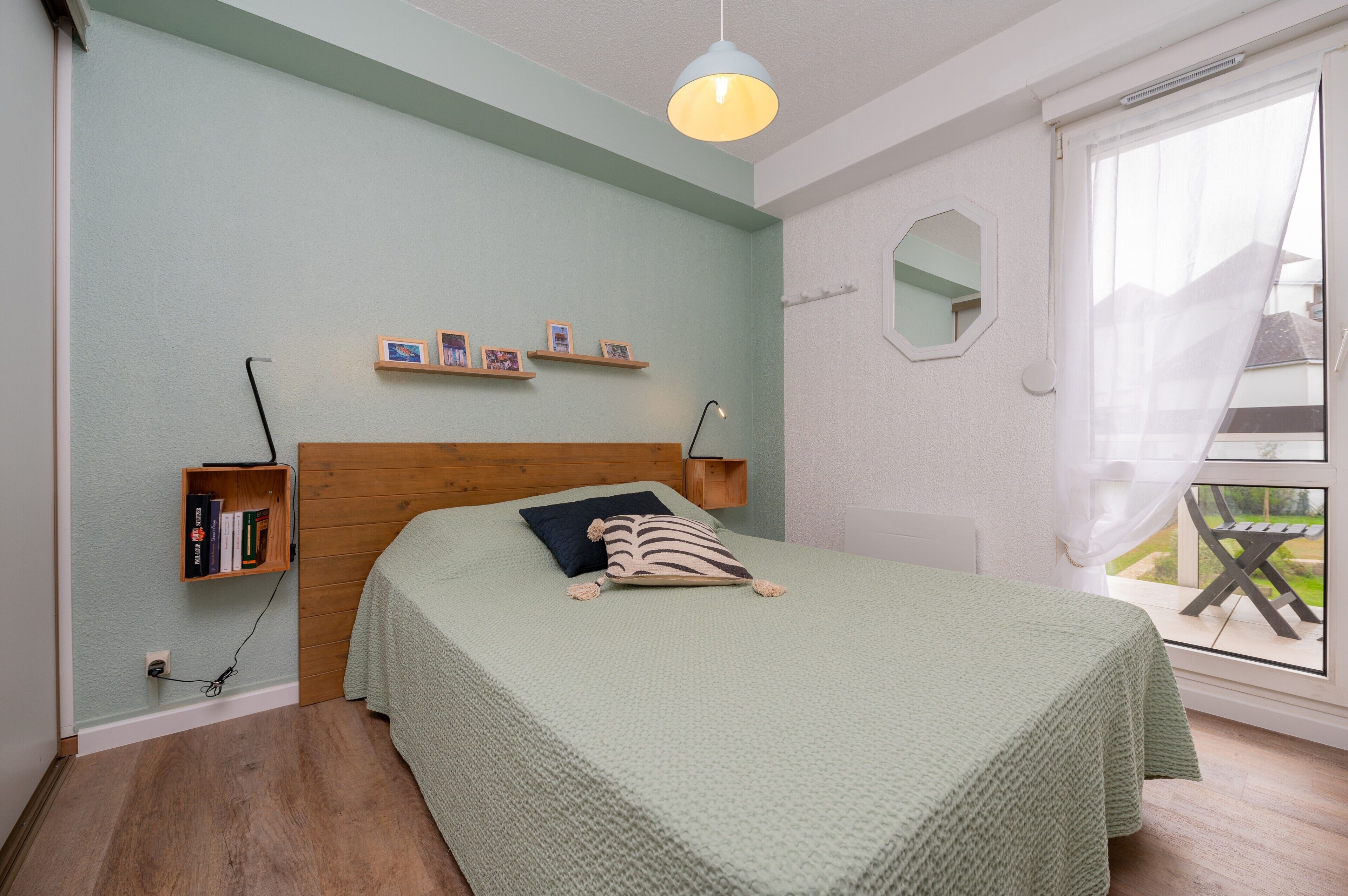 A bedroom with a bed, a wooden headboard, a shelf with books, a window with a view, and a hanging light fixture.