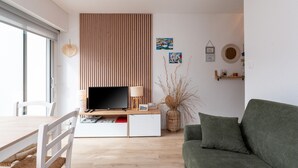 Ein modernes Wohnzimmer mit einem Flachbildfernseher, einem grünen Sofa, einer Wand mit Holzlamellen und einem Essbereich mit weißem Tisch und Stühlen.