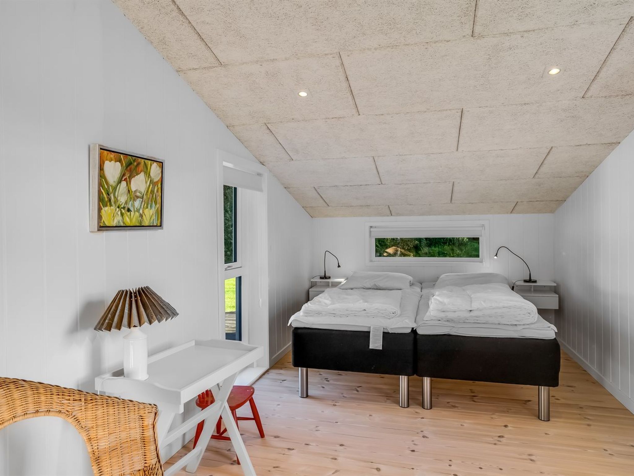 A bedroom with a double bed, a desk, a chair, and a window with a view of greenery.