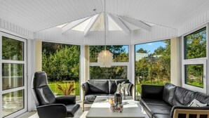 A sunroom with a modern design, featuring a black leather sofa, a black office chair, and a coffee table. Large windows provide views of a lush garden. - "Martina" - 500m from the sea by Interhome (Jerup)