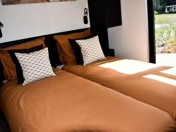 Cama doble con ropa de cama marrón, almohadas estampadas, cabecera negra y ventana con vistas a zonas verdes.