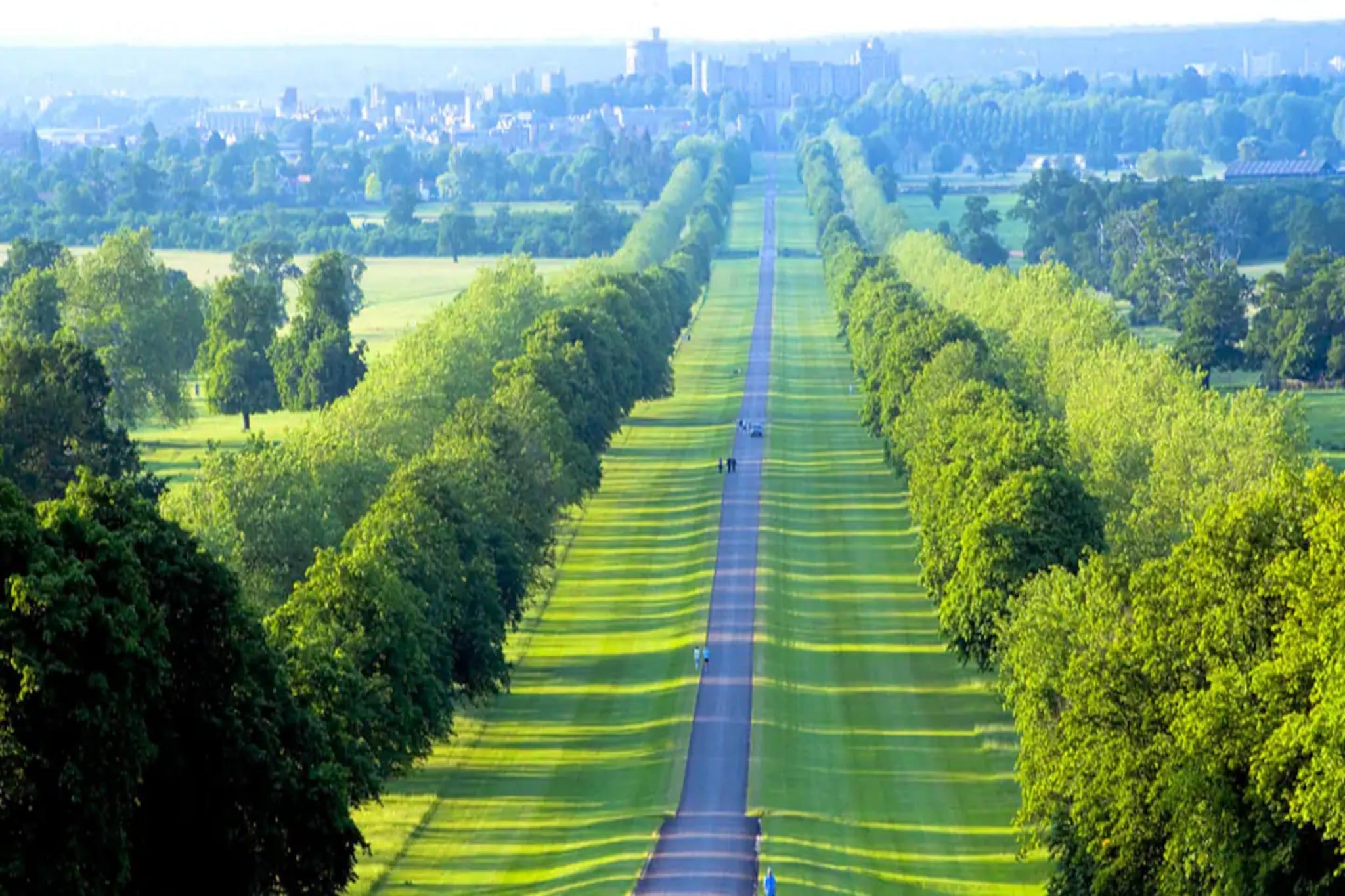 Une longue route droite, bordée de champs verdoyants et d’arbres, qui mène vers une ville lointaine à l’horizon.