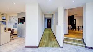 A hotel lobby with a reception desk, a waiting area with chairs, a staircase leading to a dining area, and a hallway with a patterned carpet.