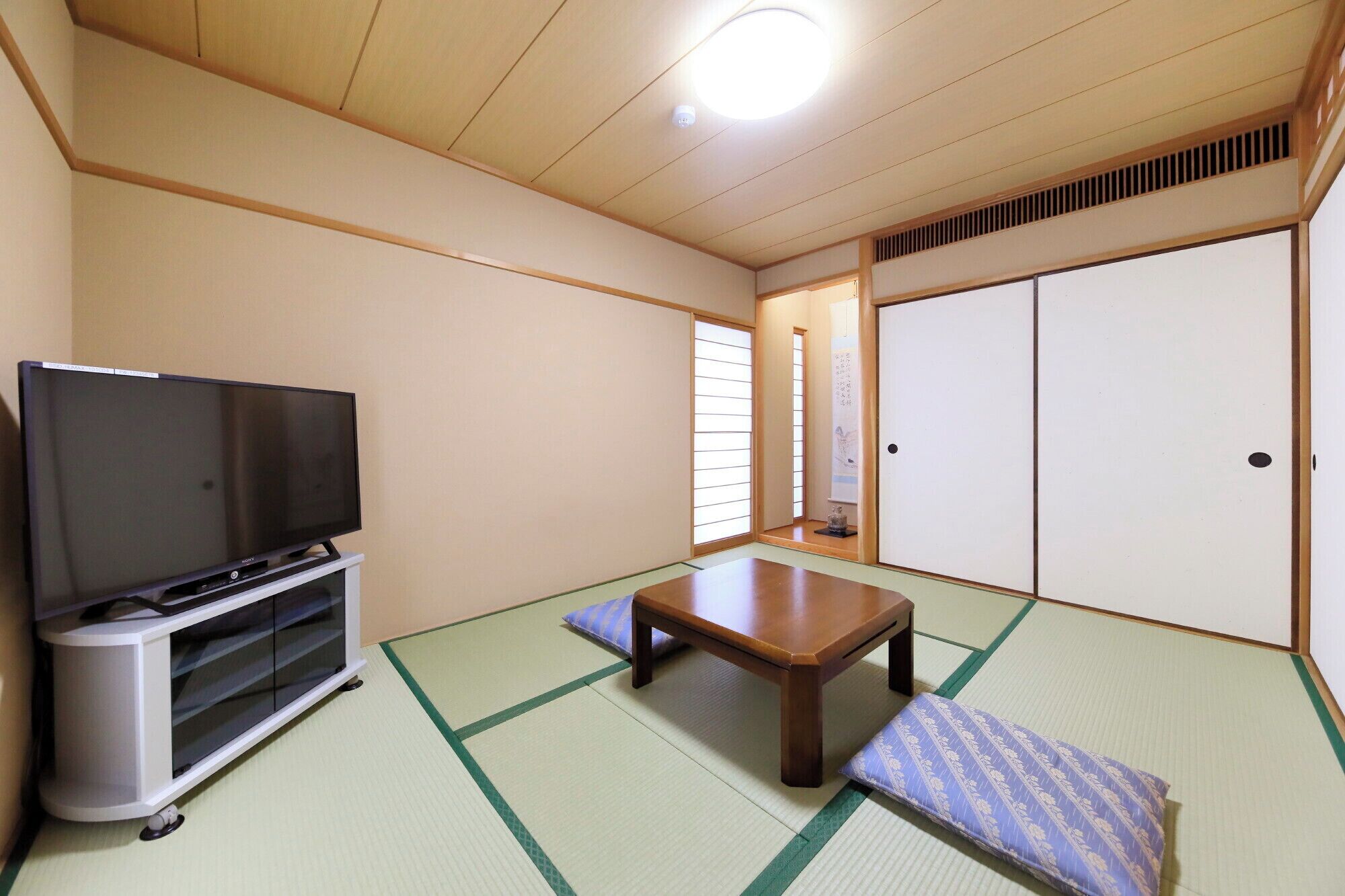 Quarto japonês tradicional com piso de tatame, televisão sobre um suporte, mesa de madeira e portas deslizantes.