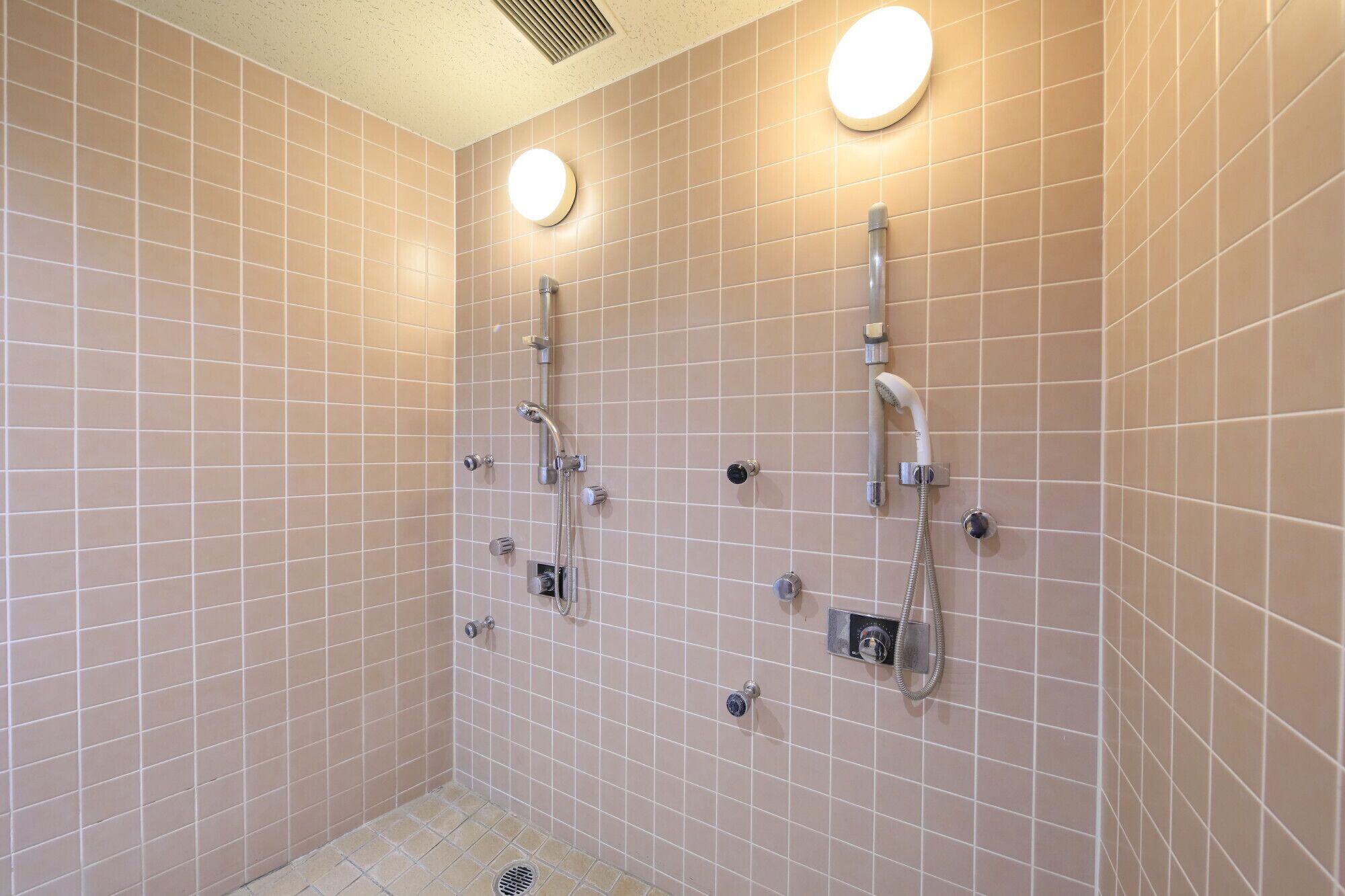 Un baño con paredes de azulejos color beige, dos cabezales de ducha y una lámpara redonda en el techo.