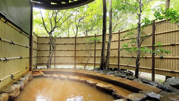 Perinteinen japanilainen onsen-kylpylä, jossa on luonnonmukainen allas, bambuseinät ja metsäinen tausta.