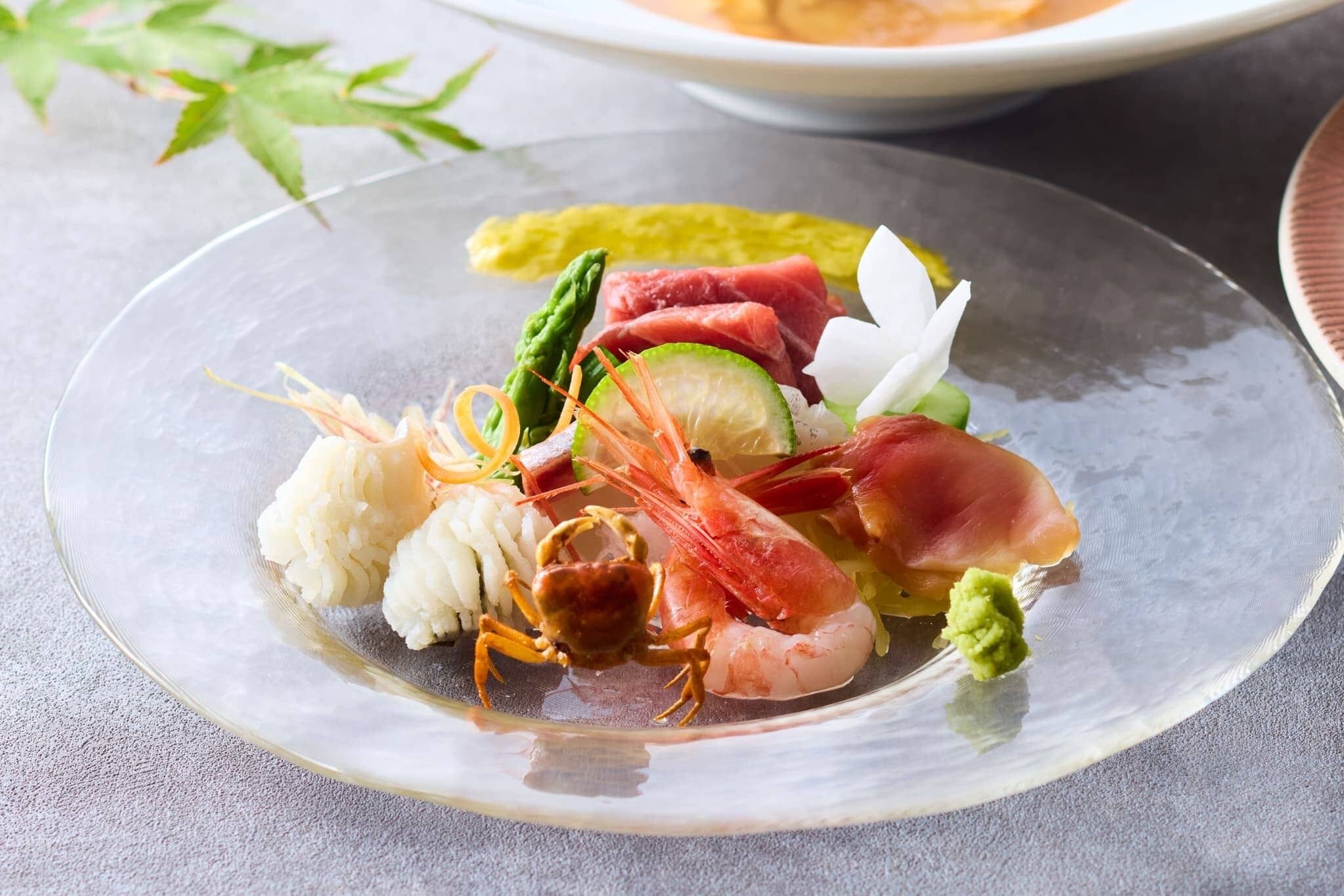 Un plato de vidrio transparente con diversos mariscos como camarones, cangrejo y filetes de pescado, acompañado de verduras y una pequeña porción de salsa verde.