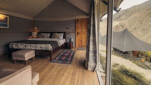 A bedroom with a large bed, a wooden bench, a bedside table, and a view of a tent outside. - Tara Mountain Sarai (Kargil)