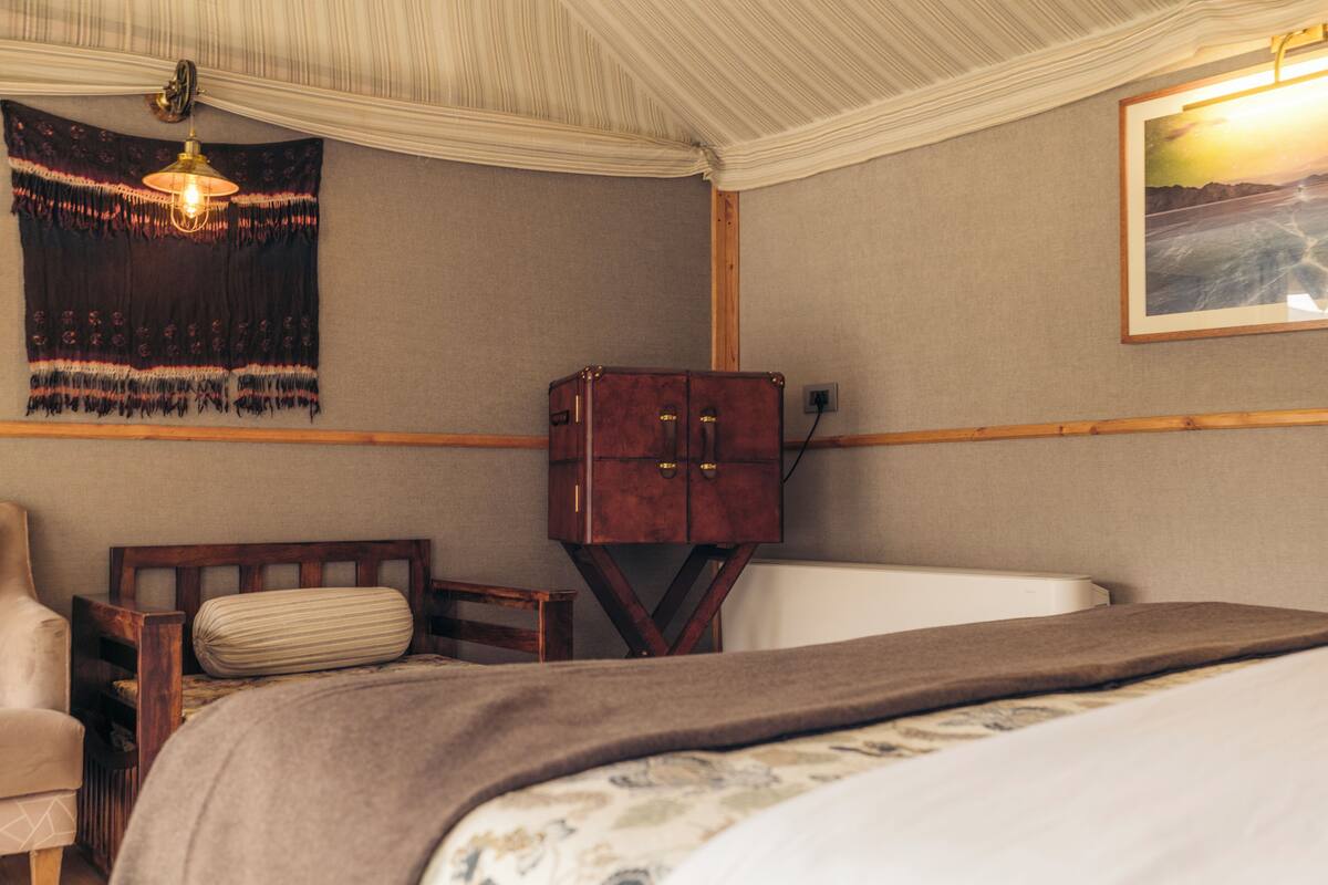 Une chambre d’hôtel avec un lit, une table de chevet en bois, une chaise, une petite armoire, une tapisserie et un tableau au mur.
