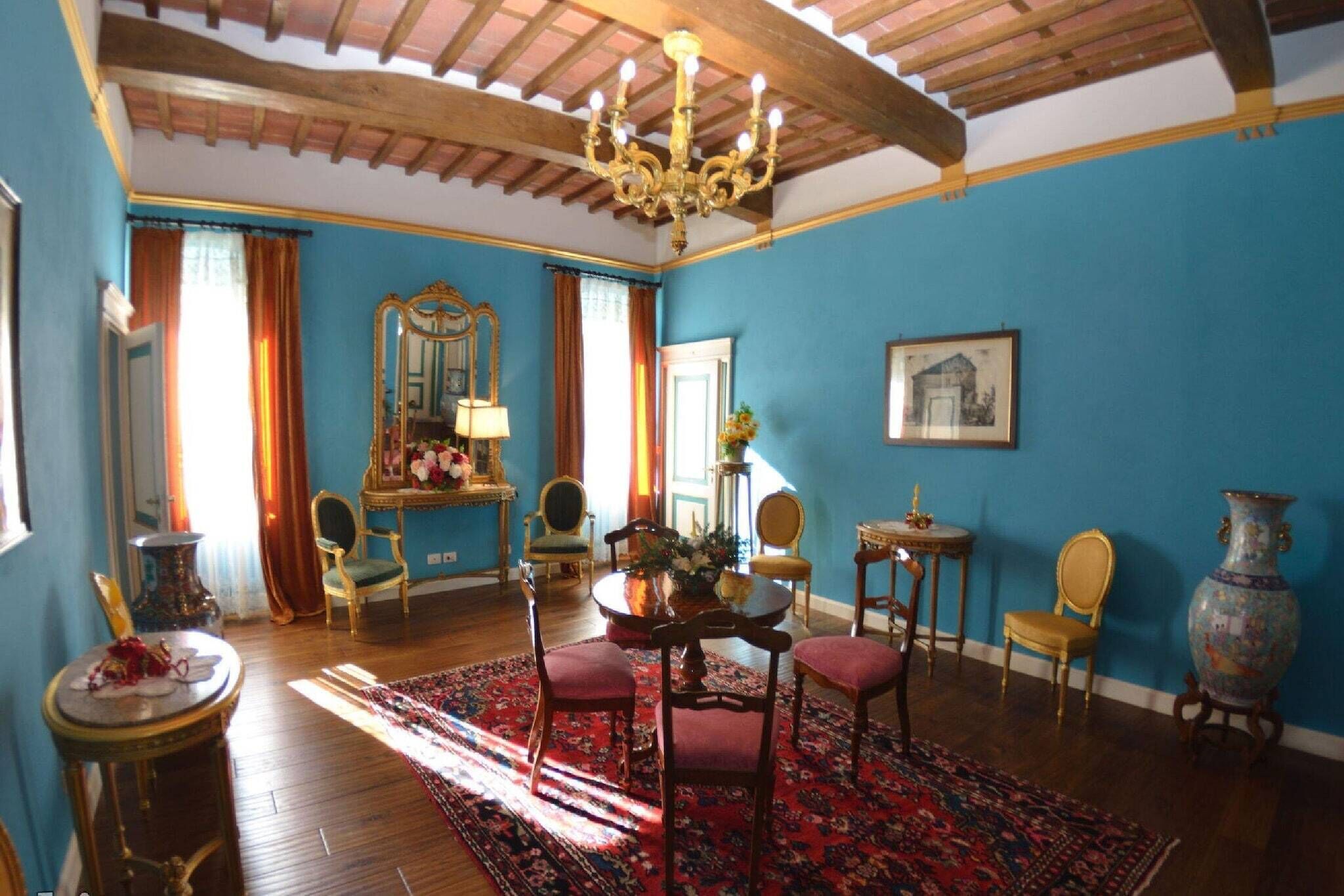 Une pièce avec un mur bleu, des poutres en bois au plafond, un lustre, une table ronde entourée de chaises, un grand miroir et un vase.