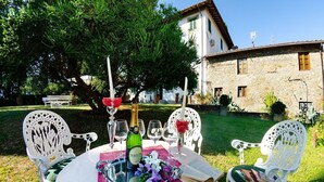Un área de comedor al aire libre con una mesa para dos, rodeada de sillas blancas, una botella de vino y un libro.