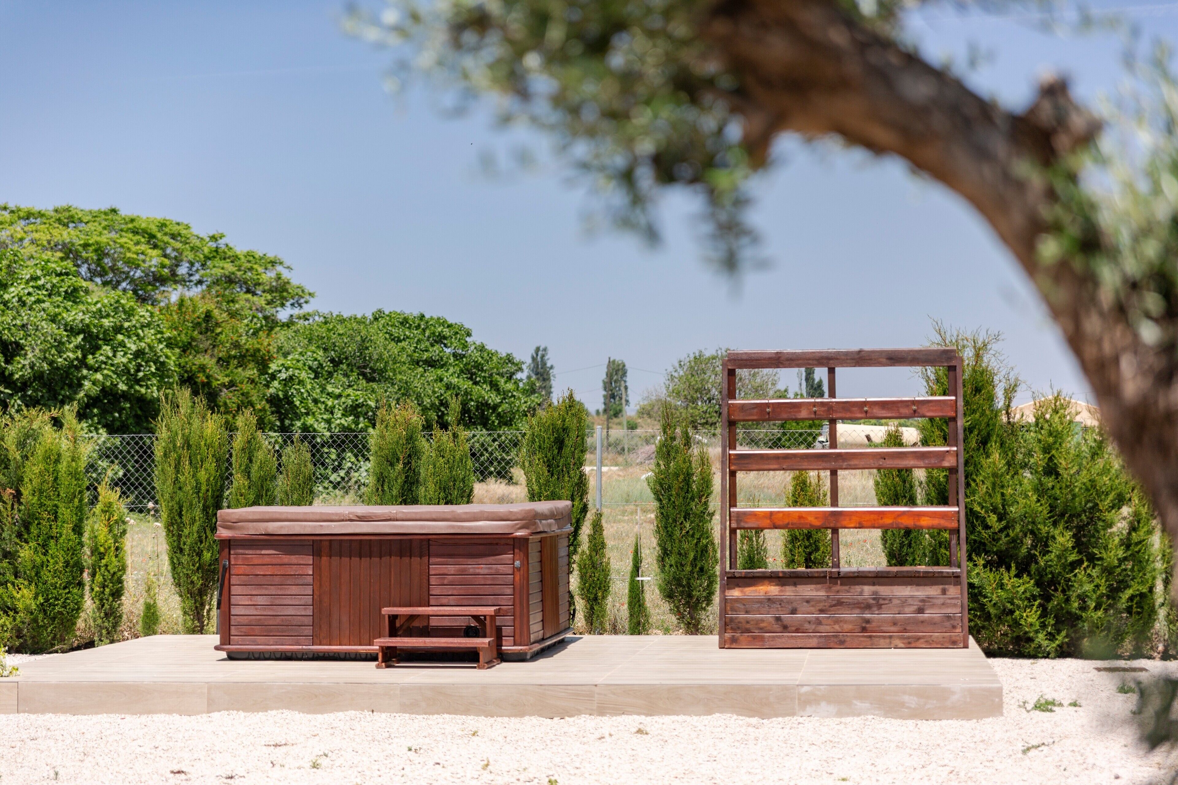 Un jacuzzi de madera y un banco de madera en un jardín con cerca y árboles.