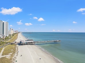 Ein Strand mit einem Pier, der sich ins Wasser erstreckt, einem sandigen Ufer und Gebäuden im Hintergrund.