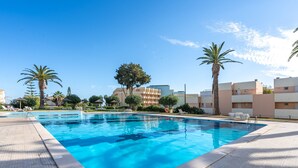 Una piscina con solárium de azulejos, palmeras y edificios modernos al fondo.