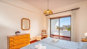 Habitación de hotel con tocador de madera, cama con ropa de cama de diseño azul, una silla, una mesita con lámpara y un amplio ventanal con vistas a edificios.