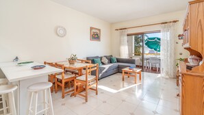 Una sala de estar moderna con un sofá, una mesa de comedor y sillas. Se observa una zona de cocina con encimera blanca y armarios de madera. Una ventana grande con cortinas es visible.