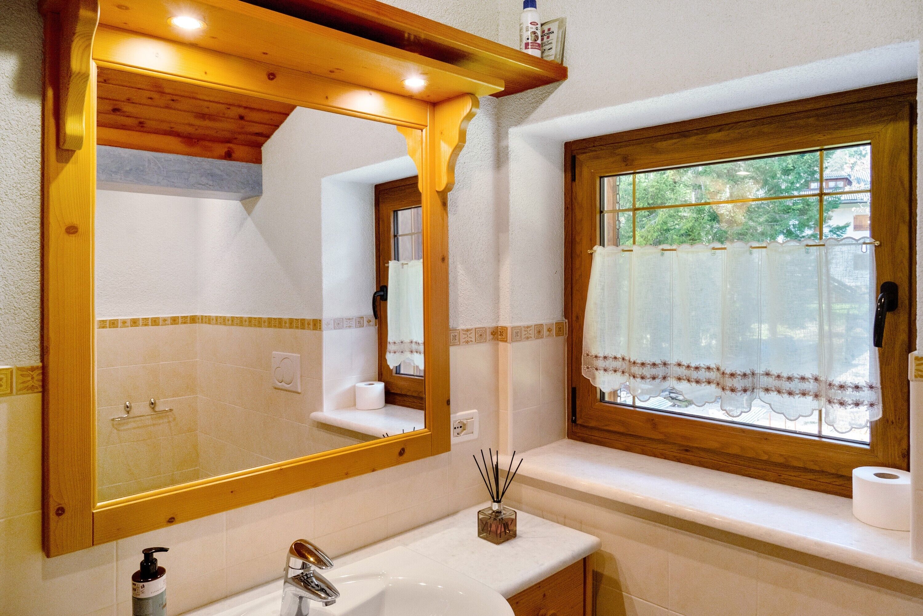 Ein Badezimmer mit einem Holzkosmetikschrank, einem großen Spiegel, einem Fenster mit Spitzgittervorhang und einer Handseife.