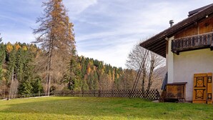 A wooden building with a balcony, a fenced yard, and a forested hill in the background. - Apartment 'Appartamento Palmina' with Mountain View, Shared Garden and Wi-Fi (Falcade)