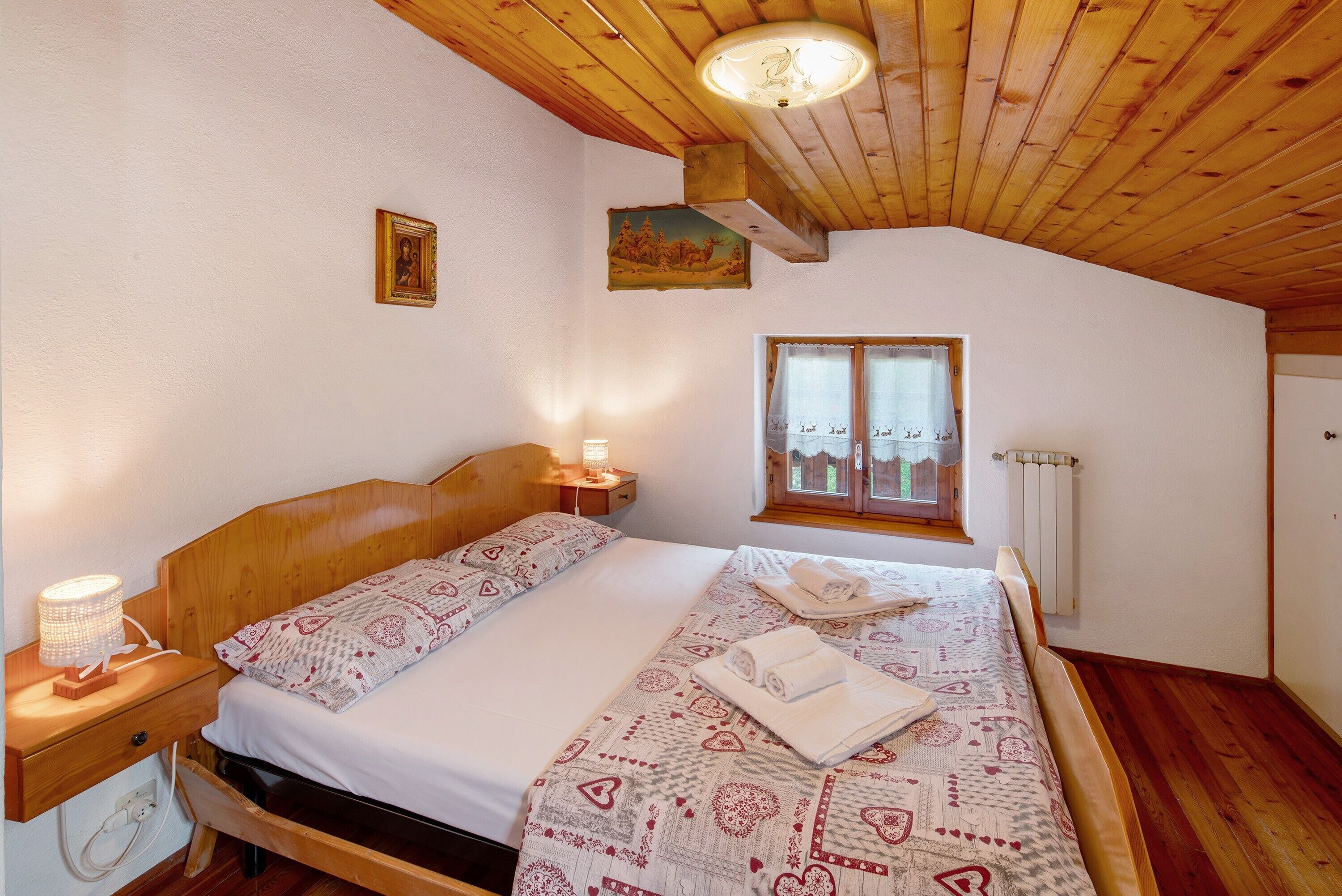A bedroom with a wooden ceiling, a bed with patterned bedding, a nightstand, a wall-mounted picture, and a window with a view.