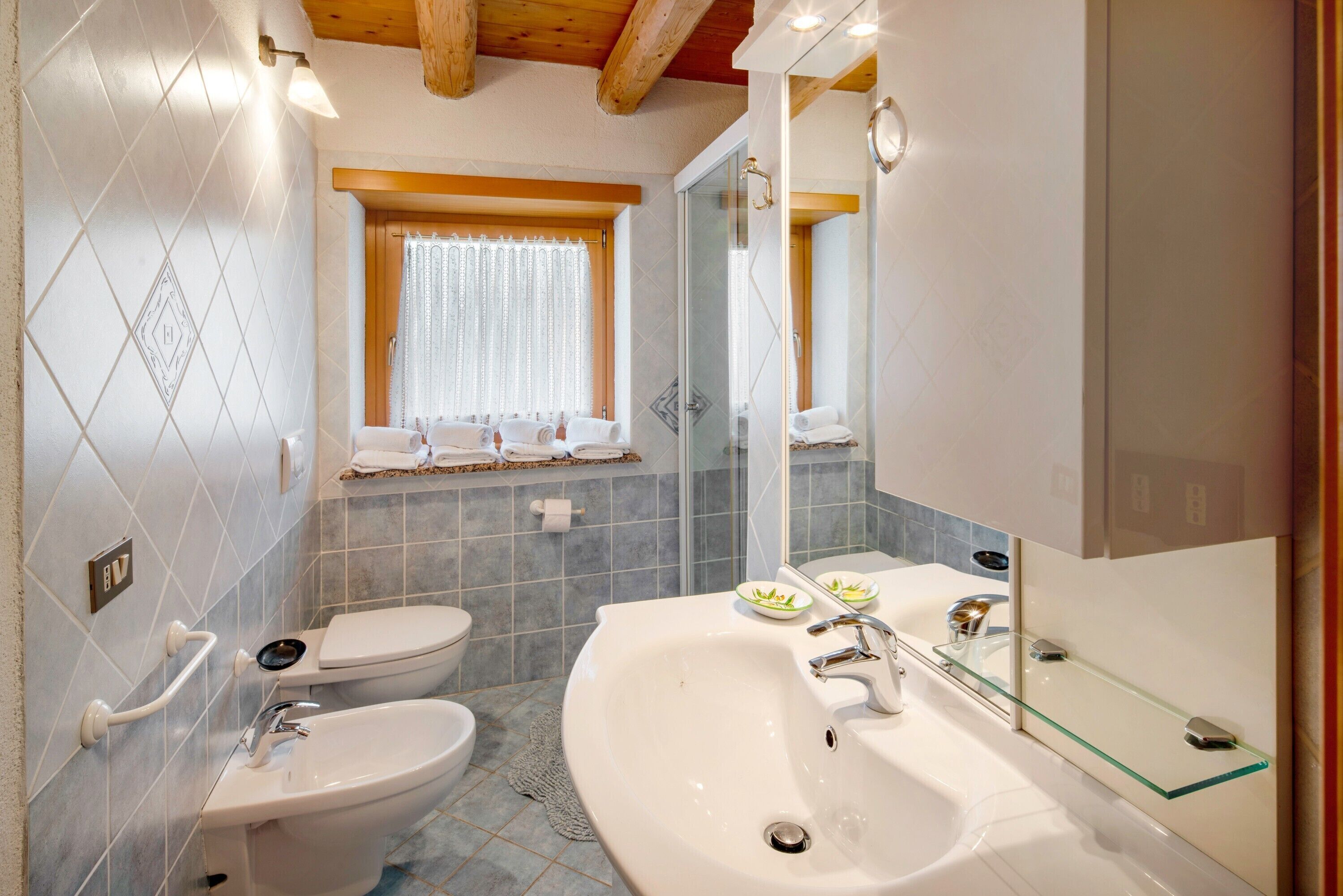 Ein Badezimmer mit weißem Waschbecken, einem Bidet, einer Toilette, einer Duschkabine aus Glas und einem Fenster mit Vorhängen.