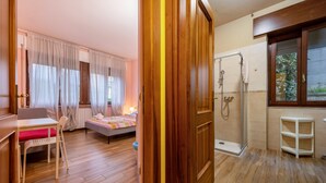 Ein Zimmer mit Holzboden, einem Bett mit gestreifter Decke, einem kleinen Tisch mit Stuhl, einer Dusche und einem Fenster mit Blick auf Grünflächen.