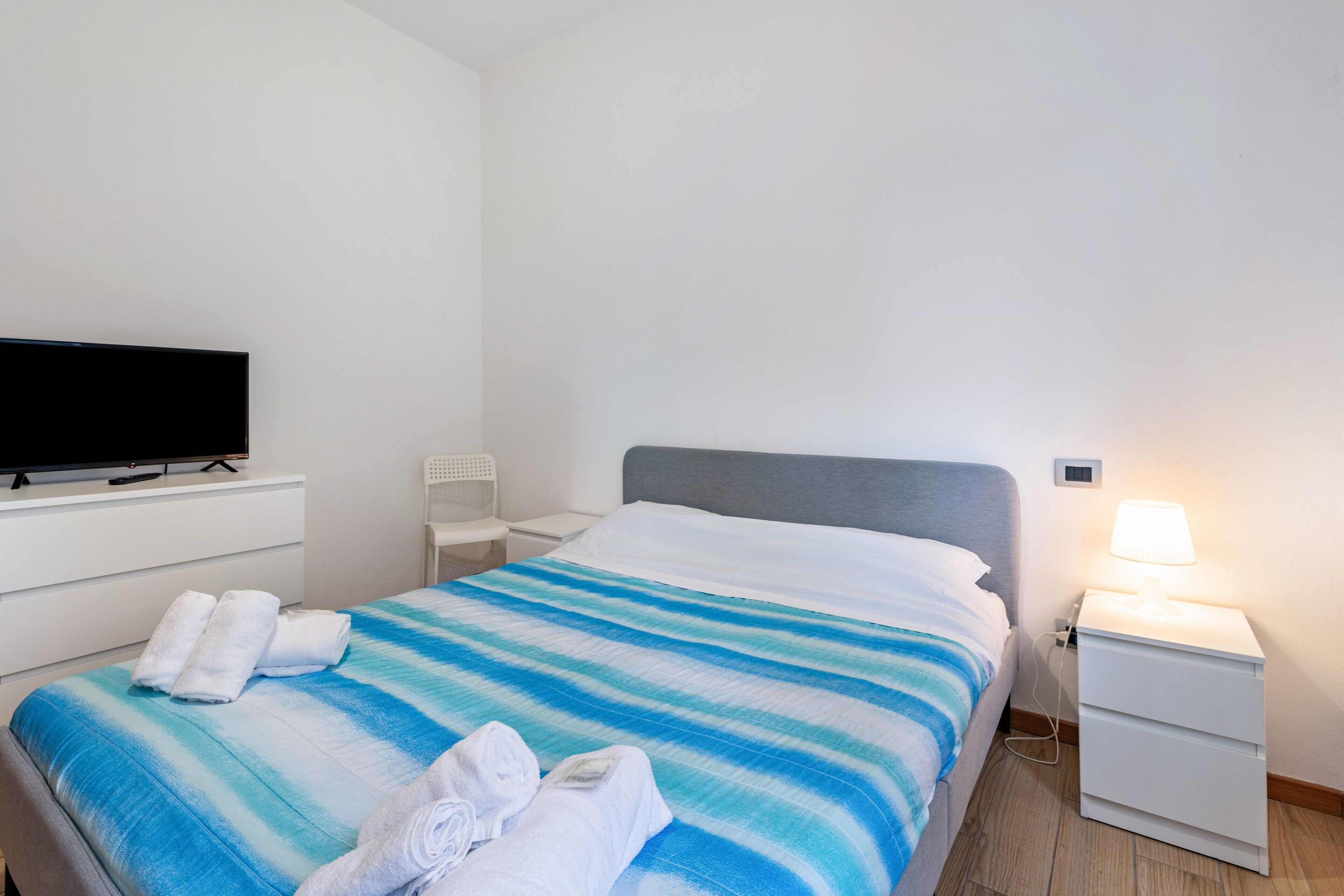 A neatly made bed with a striped bedspread, a television on a white dresser, a bedside lamp, and a small white nightstand.