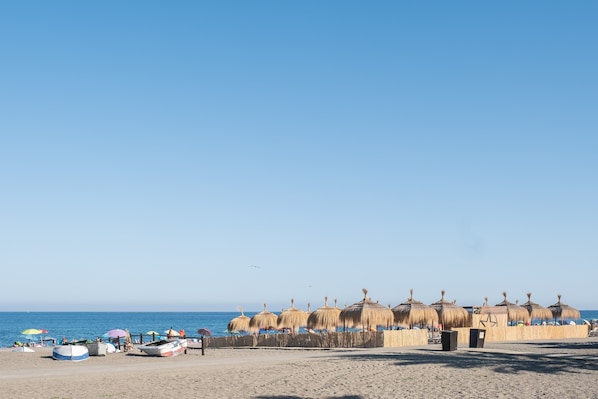 A beach with thatched umbrellas, a sandy shore, and a clear blue sky.