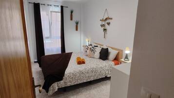 Un dormitori amb llit, una taula de nit amb lamp, una finestra amb cortines i decoració penjada a la paret.