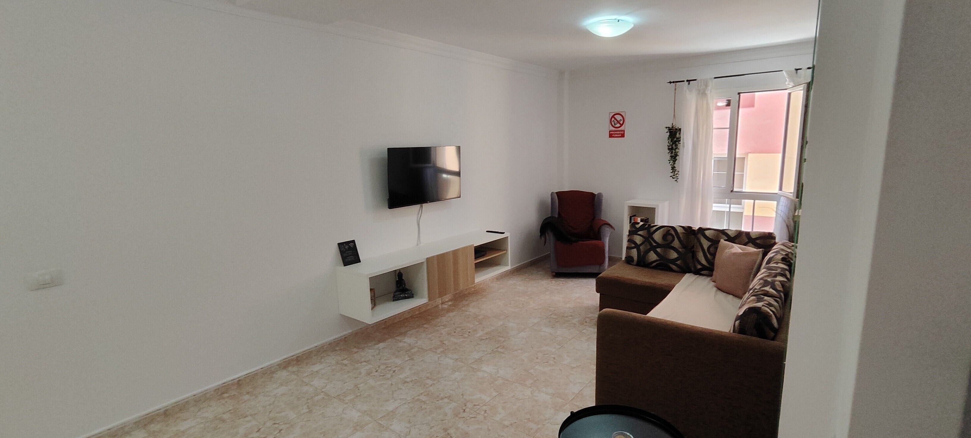 Ein Wohnzimmer mit einem an der Wand befestigten Flachbildfernseher, einem roten Sessel, einem gemusterten Sofa und einem kleinen Tisch mit einer Pflanze.