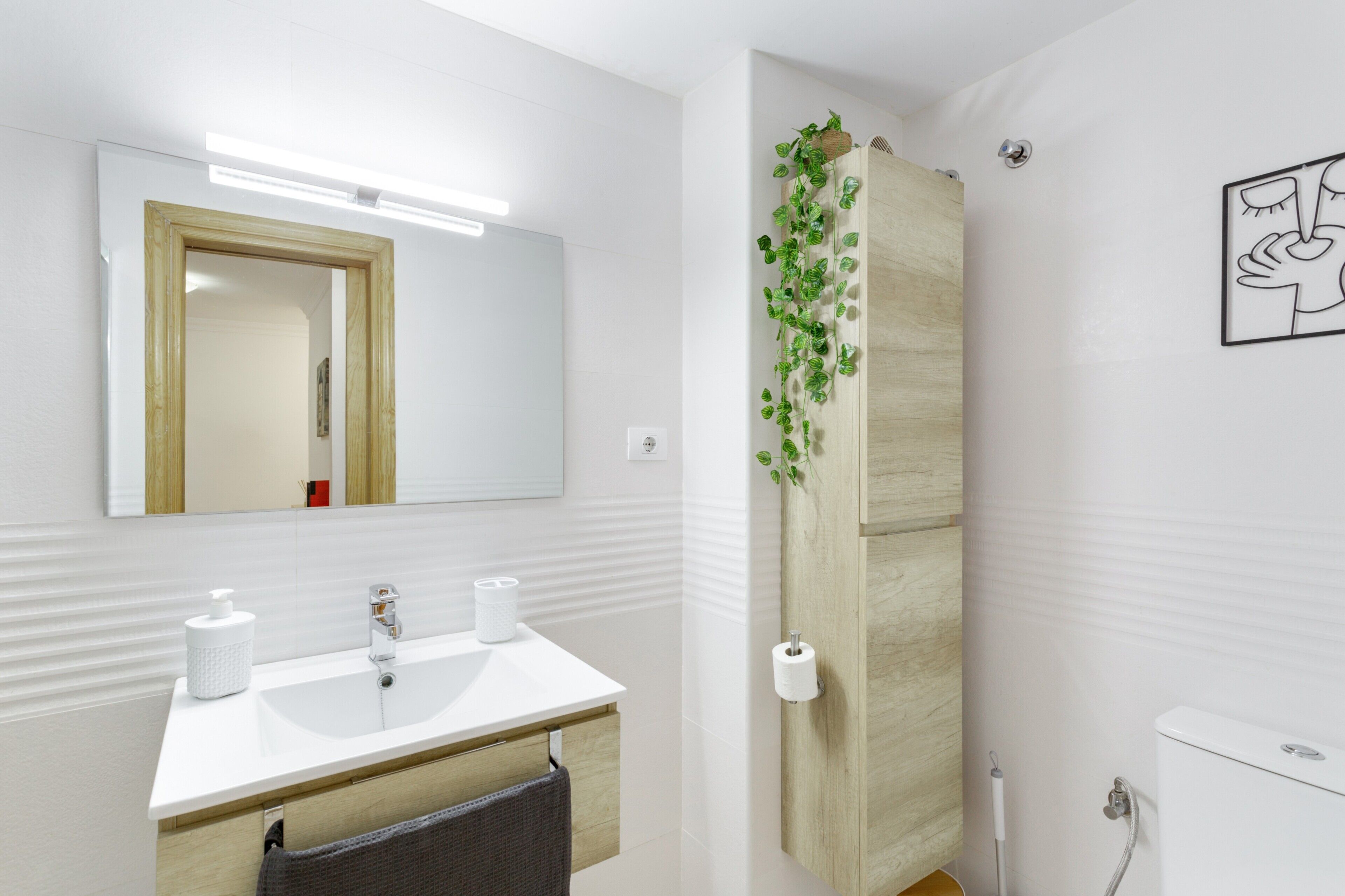 Een badkamer met een wit wastafelmeubel, spiegel en toilet. Er staat een houten kast met een groene plant erop.