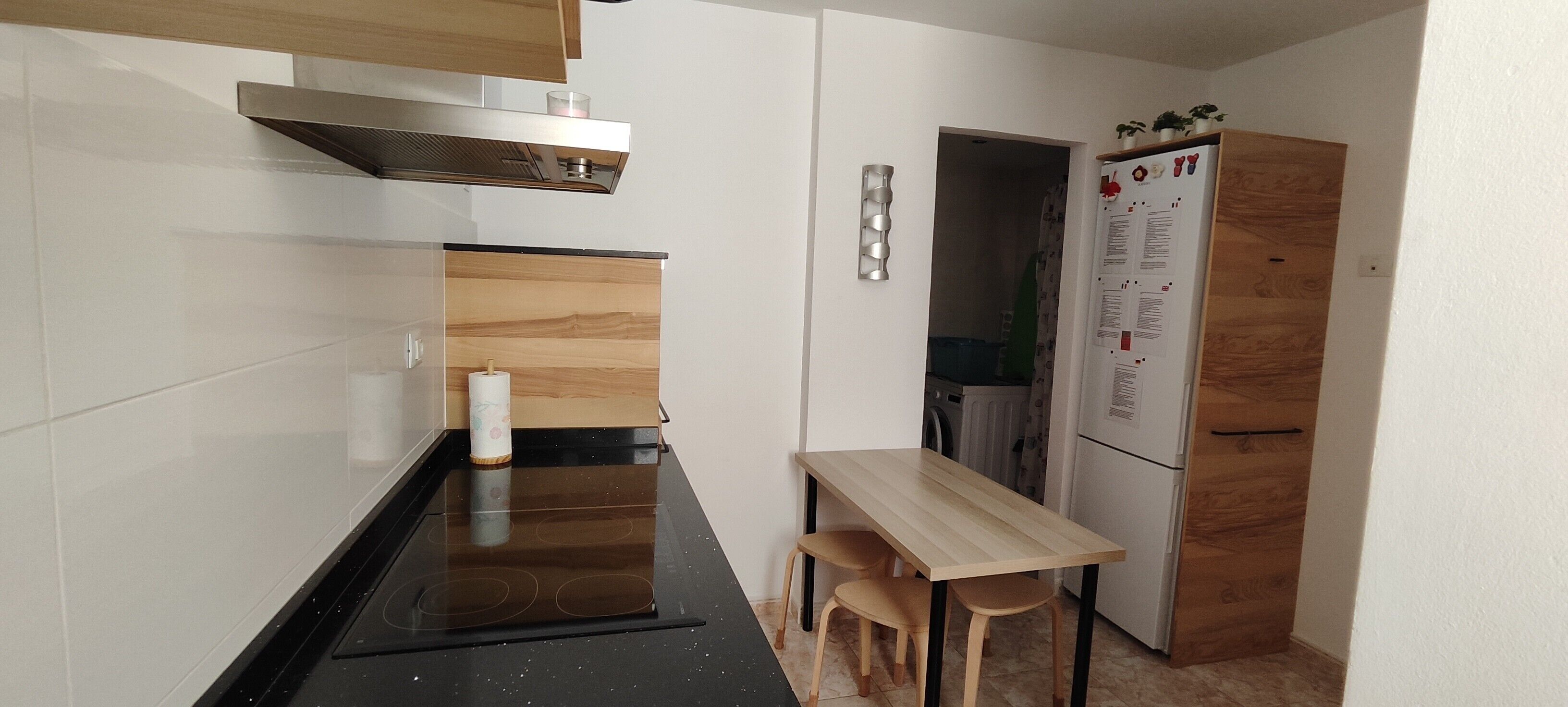 A compact kitchen with a black stove, wooden table, two stools, and a white refrigerator.