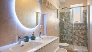 A modern bathroom with a large oval mirror, two sinks, a toilet, and a glass-enclosed shower. - Apartment 'La Casa Del Cuore' with Mountain View, Wi-Fi and Air Conditioning (Lascari)