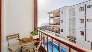 Un balcon doté d’une balustrade en bois, une table en bois ornée d’une plante, et une vue sur un immeuble blanc également doté de balcons.