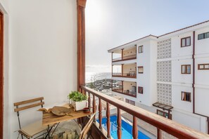 Un balcon doté d’une balustrade en bois, une table en bois ornée d’une plante, et une vue sur un immeuble blanc également doté de balcons.