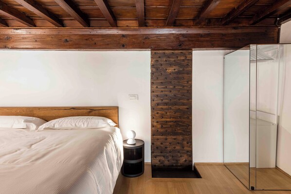 Un dormitorio con techo de madera, una cama con ropa blanca, una mesita de noche con lámpara y cabina de ducha de vidrio.