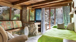 Ein rustikaler Raum mit Steinwand, Holzbalken, einem Bett mit grüner Bettwäsche, einer Bank mit floralen Kissen und einem Fenster mit gemusterten Vorhängen.