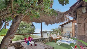 Eine Holzterrasse mit Tisch und Stühlen, einem Reetdach und einer Steinmauer.