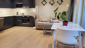 A modern kitchen with dark cabinets, a microwave, and a built-in oven. There is a dining area with a white table and chairs, a sofa with cushions, and a potted plant. - Oslo Center, Norway, your second home. (Oslo)