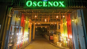 The entrance to a hotel named 'OSCENOX' with a neon sign, a wooden archway, hanging lights, and a signboard for a laundry service.