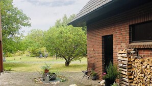 En byggnad av tegel med en stenlagd uteplats, en hund som sitter på förgrunden och en hög med vedstaplar till höger.