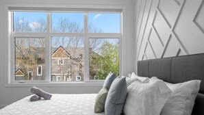 A bedroom with a large window, a bed with pillows, and a view of a house.