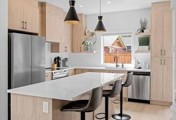 Una cocina moderna con una isla central, taburetes de bar, iluminación colgante y vistas a una casa.