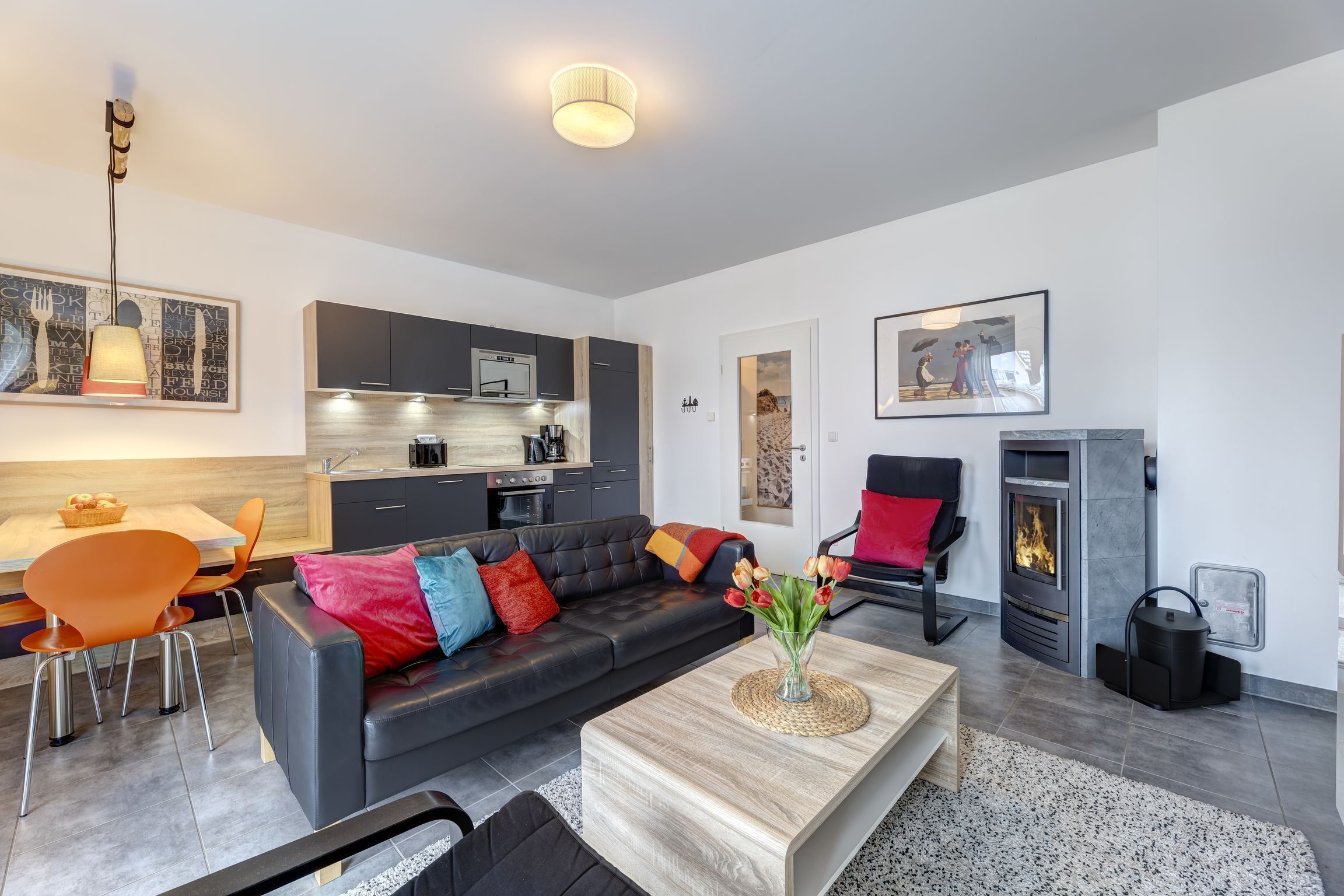 Un salon moderne avec un canapé, une table basse et une cheminée. On peut apercevoir une cuisine avec des meubles sombres et un coin repas avec des chaises orange.